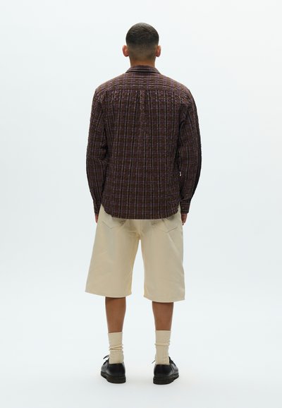 Φαρδιά πουκάμισα σε μοβ και καφέ καρό ύφασμα, σορτς σε ανοιχτό μπεζ, συνδυασμένα με σκούρα παπούτσια και κρεμ κάλτσες.