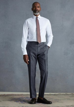 Weißes Langarmhemd kombiniert mit dunkelgrauen karierten Hosen und schwarzen Slippern. Das Outfit vermittelt ein maßgeschneidertes, professionelles Erscheinungsbild.
