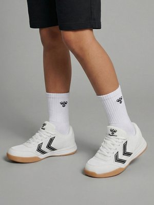 Nogi w białych sportowych butach ze wzorem czarnych pasków chevron i białych skarpetach za kostkę z czarnym logo pszczoły, stojące na szarej podłodze.