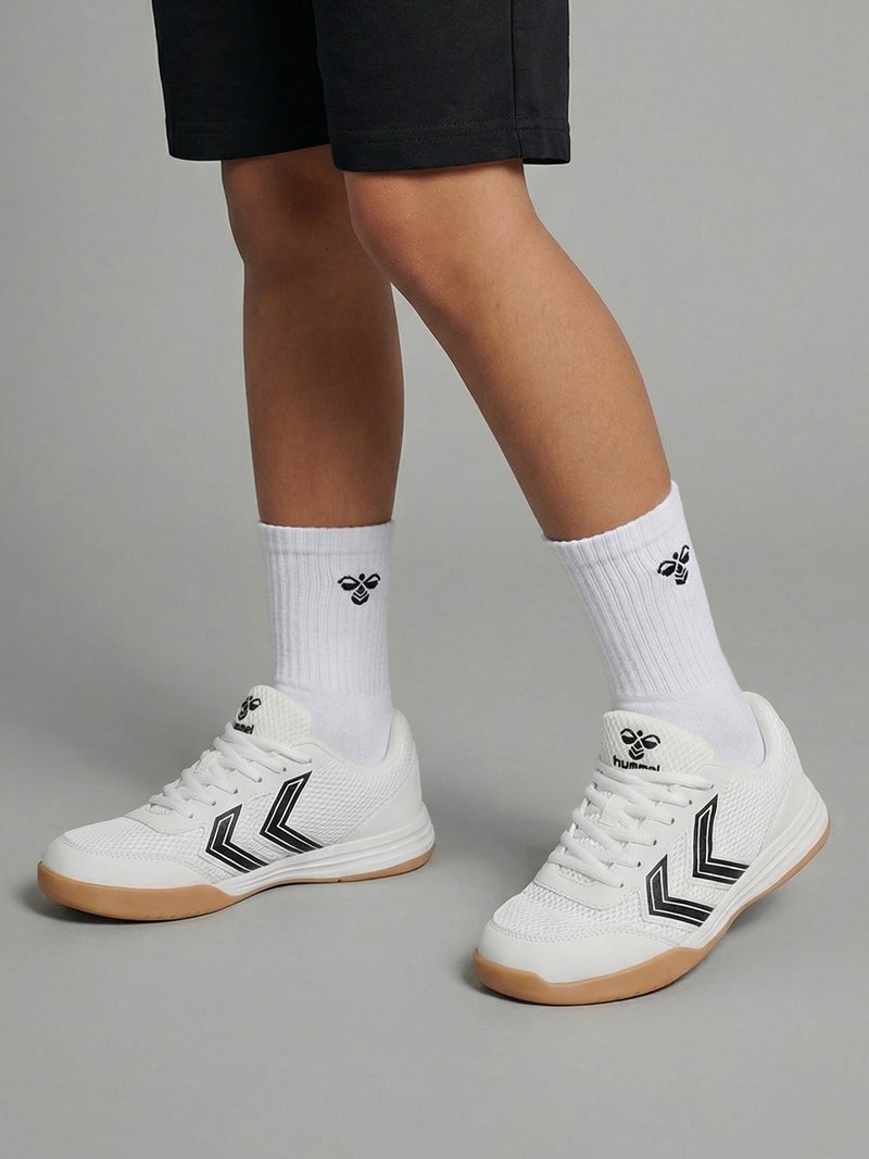 Benen met witte sportschoenen met zwarte chevronstrepen en witte kousen met een zwart bijlogo, staand op een grijze vloer.