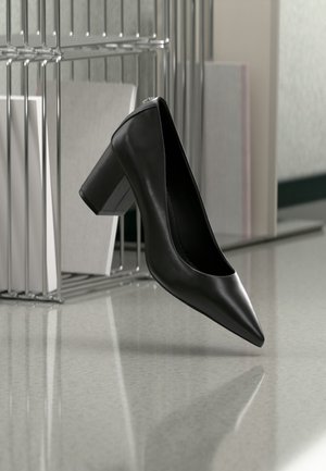 Pantof cu toc înalt negru din piele, cu vârf ascuțit și toc gros, suspendat deasupra unei podele reflectorizante, lângă un organizator metalic pentru dosare cu documente.