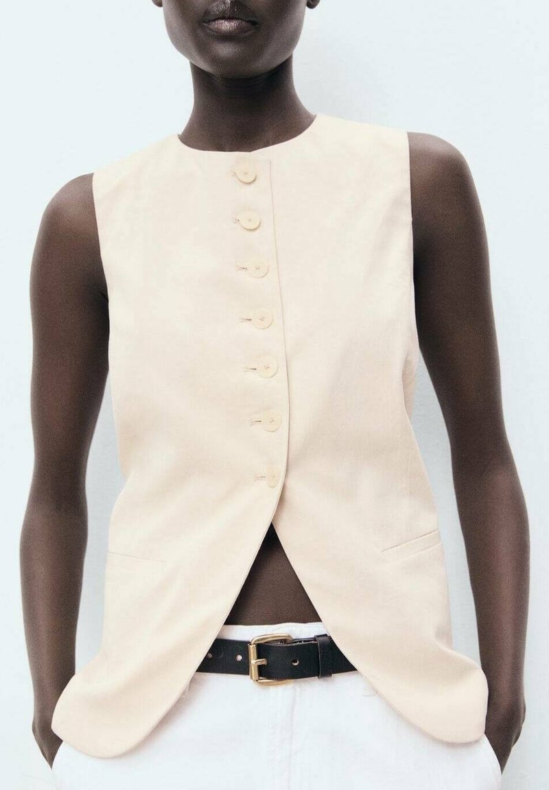Beige ärmelloses Oberteil mit geradem Schnitt, ausgestattet mit einer Reihe von sechs Knöpfen und seitlichen Taschen, getragen mit einem schwarzen Gürtel über weißen Hosen.