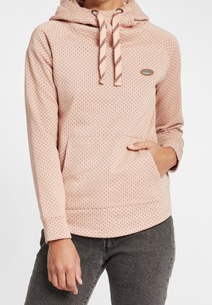 Sweat à capuche rose clair avec petits points marron, capuche avec cordon ajustable, poche avant et écusson de logo ovale sur la poitrine, porté avec un jean foncé.