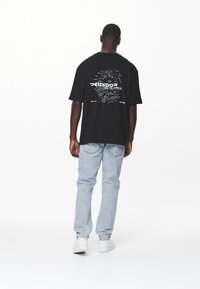 T-shirt di cotone nero con maniche corte, con una stampa grafica sulla schiena, abbinata a jeans di denim blu chiaro e sneakers bianche.