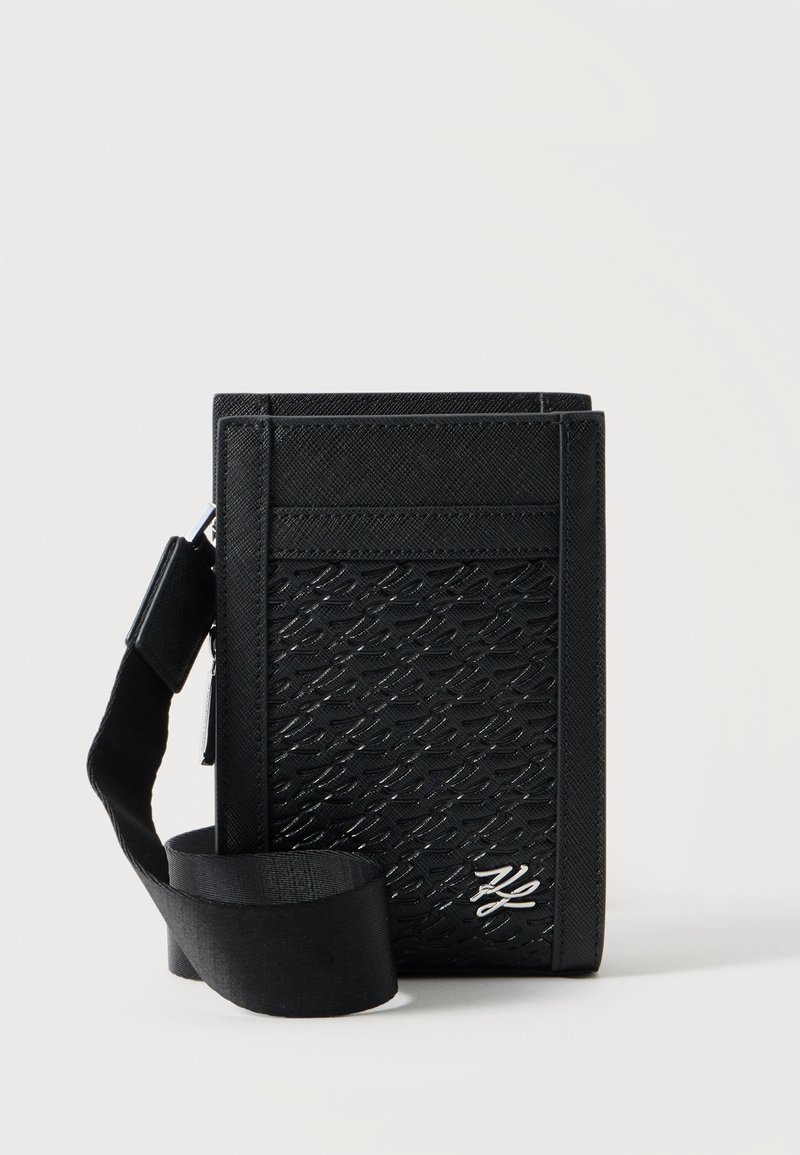 Μαύρο υφασμάτινο τσαντάκι crossbody με ρυθμιζόμενο υφασμάτινο ιμάντα, ανάγλυφο σχέδιο και ασημένιο λογότυπο στην μπροστινή όψη σε απλό φόντο.