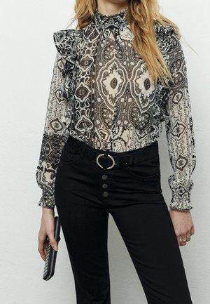 Femme en blouse à col montant noire et blanche avec des volants, pantalon taille haute noir à boutons apparents, tenant une petite pochette.