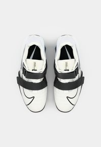 Baskets de sport blanches avec des accents et des sangles noires, tige texturée, design incurvé et logo visible sur le côté.