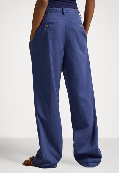 Granatowe spodnie o szerokich nogawkach mają płaskie przody, boczne kieszenie, tylne kieszenie zapinane na guziki oraz gładką fakturę tkaniny.