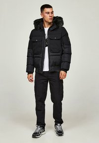 Casaco acolchoado preto com capuz de pelo sintético, dois bolsos frontais, fecho de correr, usado sobre uma t-shirt branca com estampa gráfica, combinado com calças cargo pretas e ténis.