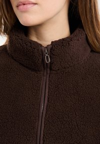 Brązowa kurtka polarowa z wysokim kołnierzem i zamkiem z przodu. Tkanina ma teksturę o miękkim wyglądzie, podkreślającą ciepło i wygodę.