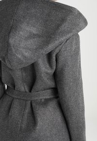 Abrigo de lana gris con una gran capucha, detalle de cintura ajustada y una textura suave. La vista posterior muestra un diseño plisado y un cierre de cinturón.