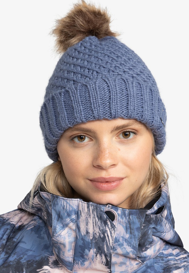 Gorro de punto azul con un patrón texturizado y un pompón de piel marrón en la parte superior, llevado con una chaqueta de colores y patrones.