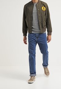 Veste bomber vert olive avec col et poignets côtelés, pull en maille gris en dessous, jean en denim bleu et baskets marron clair.