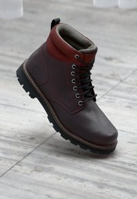 Dark brown leather hiking boot with red padded collar, black laces, and rugged black sole on light gray marble floor.