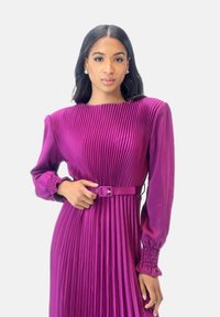 Robe plissée violette à manches longues, avec des poignets froncés et une ceinture assortie à la taille. Texture satinée lisse avec des lignes verticales.