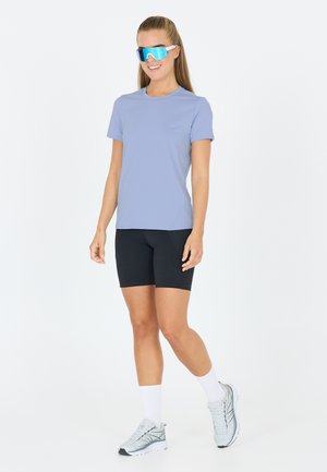 Maglietta sportiva azzurro chiaro a maniche corte, abbinata a pantaloni ciclismo neri e scarpe da ginnastica grigie. Grandi occhiali da sole specchiati completano il look.