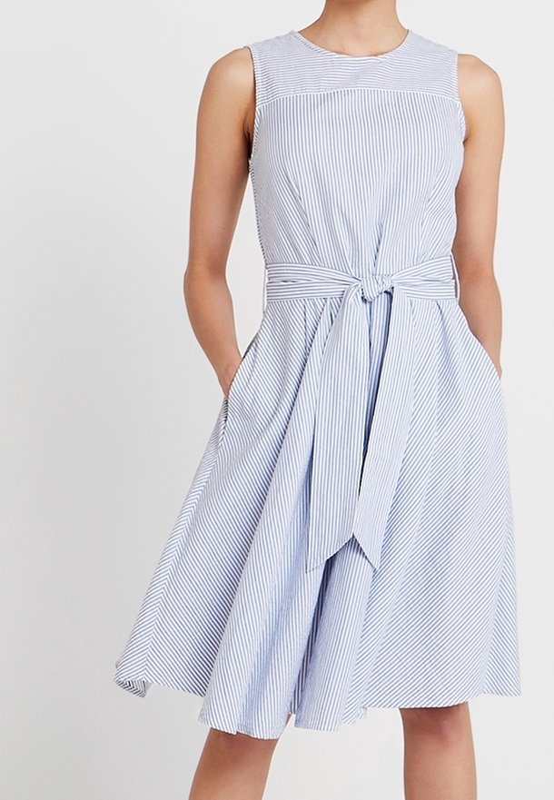 Robe sans manches rayée bleu clair et blanc avec ceinture nouée à la taille et jupe évasée longueur genoux, portée avec les mains dans les poches.