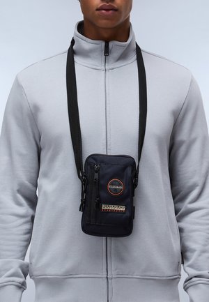 Zwarte nylon schoudertas met een rits sluiting, voorzien van een cirkelvormige logo-patch en een verstelbare strap, gedragen over een lichtgrijs ritsvest.