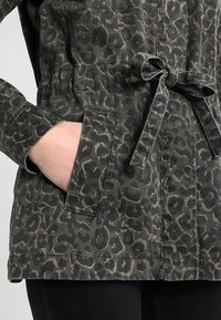 Chaqueta de estampado de leopardo en tonos verdes y marrones, con cintura ceñida con cinturón, dos bolsillos laterales y una tela suave y texturizada.