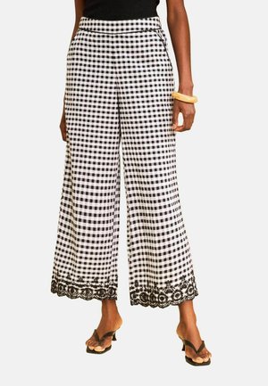 Nainen, joka käyttää mustavalkoruudullisia leveälahkeisia housuja, joissa on musta kukallinen pitsireunus lahkeissa, mustat sandaalit ja keltainen rannekoru.