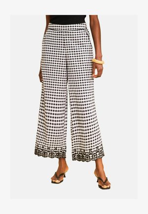 Femeie care poartă pantaloni largi cu carouri alb-negru cu dantelă florală neagră la tiv, sandale negre și o brățară galbenă.