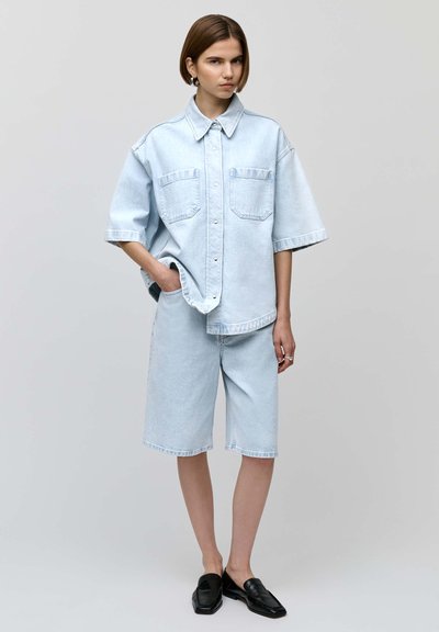 Camisa de mezclilla oversized de color azul claro con dos bolsillos en el pecho, combinada con pantalones cortos de mezclilla hasta la rodilla, llevada con zapatos negros sin cordones.