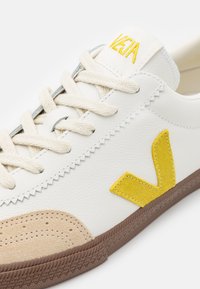 Fehér bőr sneaker sárga velúr logóval és bézs velúr lábujjvédővel. Kerek fűzőkkel és texturált barna gumitalppal rendelkezik.