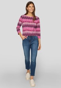 Langärmliges Oberteil mit pink-grauem Chevron-Muster, kombiniert mit blauen Jeans und weißen Slippern, Model mit langen braunen Haaren steht selbstbewusst.