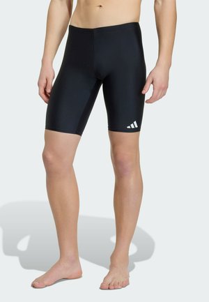Męski tors i nogi w czarnych krótkich kąpielówkach do kolan z białym logo Adidas na lewej udzie, stojące boso na białym tle.