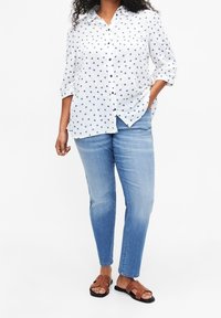 White button-up shirt with navy heart pattern, rolled sleeves, paired with light blue jeans and brown sandals with stud accents.