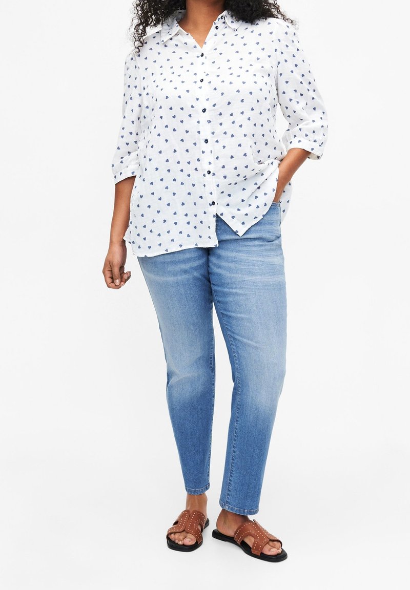White button-up shirt with navy heart pattern, rolled sleeves, paired with light blue jeans and brown sandals with stud accents.