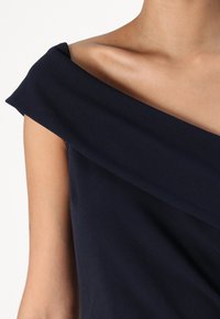 Navy off-shoulder kjole med drapert hals og glatt tekstur. Nærbilde viser mykt stoff og rene linjer.