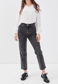 Femme portant une blouse blanche à manches longues avec des poignets smockés, un jean noir délavé taille haute et des chaussures à lacets noires et brillantes, se tenant sur un sol blanc.