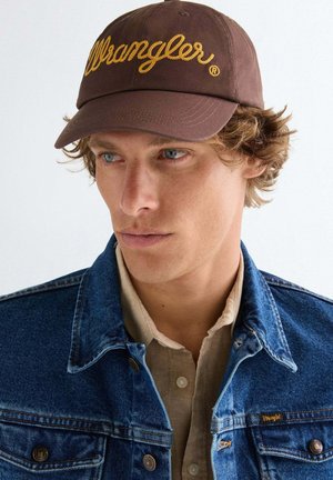 Junger Mann mit blauen Augen, der eine braune Wrangler-Kappe, ein beiges Hemd zum Knöpfen und eine blaue Denimjacke mit Wrangler-Logo trägt.