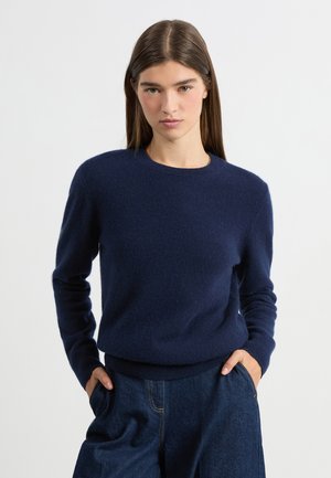 Femme aux longs cheveux bruns portant un pull bleu marine et un pantalon en jean, les mains dans les poches, sur un fond blanc uni.