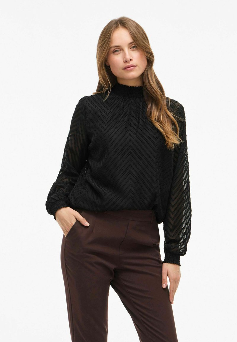 Femme aux longs cheveux portant un haut noir texturé à manches longues et un pantalon marron foncé, debout avec une main dans la poche sur fond blanc.