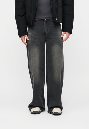 Άτομο που φοράει φαρδιά ξεθωριασμένα μαύρα τζιν, συνδυασμένα με λευκά και μαύρα αθλητικά παπούτσια, μαύρο μπουφάν και σκούρο γκρι τοπ.