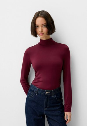 Burgundy long-sleeve bodysuit with a high neck; fitted design in smooth fabric. Paired with dark denim jeans featuring front pockets.