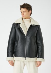 Giubbotto di pelle nera con collo e polsini in shearling, dotato di zip frontale e tasche laterali. Materiale testurizzato e cinturini regolabili.