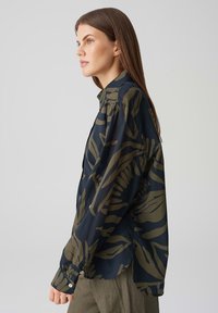 Camicia button-up di un blu navy scuro con grandi motivi a foglia verde oliva. Presenta un colletto, maniche lunghe e una vestibilità comoda. Tessuto dal tocco morbido.