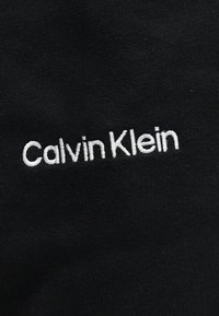 Μαύρο ύφασμα με λευκό κεντητό κείμενο που γράφει «Calvin Klein», ελαφρώς κεκλιμένο.