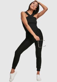 Schwarzes ärmelloses Oberteil mit Kreuzträgern, kombiniert mit schwarzen Spitzen-Jogginghosen und weißen Sneakers, die über strukturierte Muster und elastische Bündchen verfügen.