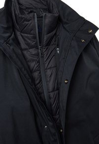 Chaqueta negra con forro interior acolchado, cuello alto y múltiples bolsillos con cremallera y detalles en latón. Textura de tela suave en toda la prenda.