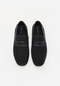 Chaussures noires à enfiler en matériau mesh, avec des accents en cuir lisse, bout rond, et un design subtil sur la sangle. Semelle plate, détails minimaux.