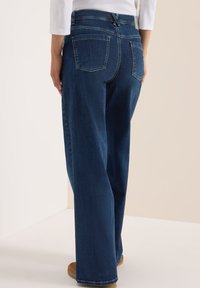 Jean en denim bleu à jambe large avec une taille mi-haute, poches arrière visibles et un motif de couture classique. Le matériau présente une légère texture.