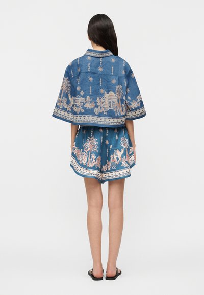 Γυναίκα με μακριά σκουρόχρωμα μαλλιά που φοράει μπλε cropped τζάκετ με κοντό μανίκι και ταιριαστά σορτς με μπεζ λεπτομερή τοπικό σχέδιο, στέκεται γυρισμένη με την πλάτη.