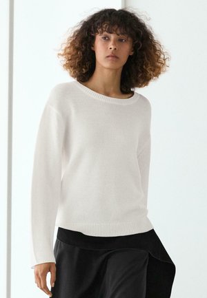 Kvinde med krøllet hår iført en hvid striktrøje og sort nederdel, stående mod en ensfarvet lys baggrund.