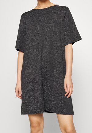 Frau trägt ein lockeres, kurzärmeliges schwarzes Kleid mit einem dezenten silbernen Punktmuster und steht vor einem schlichten, hellen Hintergrund.