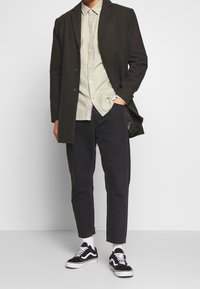 Muž v černém kabátu, světle béžové košili na knoflíky, černých zkrácených kalhotách, bílých ponožkách a černých teniskách s bílými tkaničkami a pruhem.