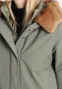 Olivově zelená bunda se zipovým zapínáním, vybavená odnímatelným béžovým límcem z umělé kožešiny a hladkou texturou.